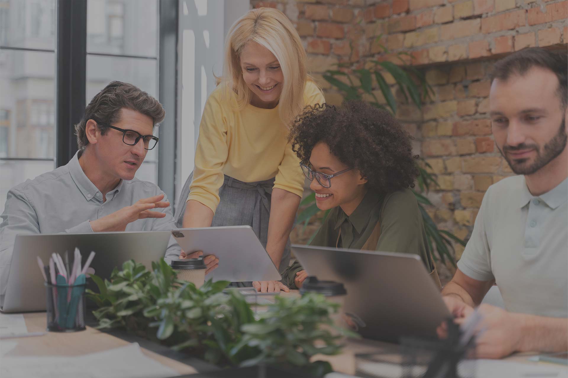 A diverse group of four people is engaged in a collaborative discussion around a table with laptops and digital tablets. The text "AGILE CULTURE – Flexibler Umgang mit Veränderungen" is prominently displayed, promoting agile work culture. One person is explaining something, while others listen attentively and smile. The background includes a brick wall and some plants, giving the setting a modern, creative workspace vibe.