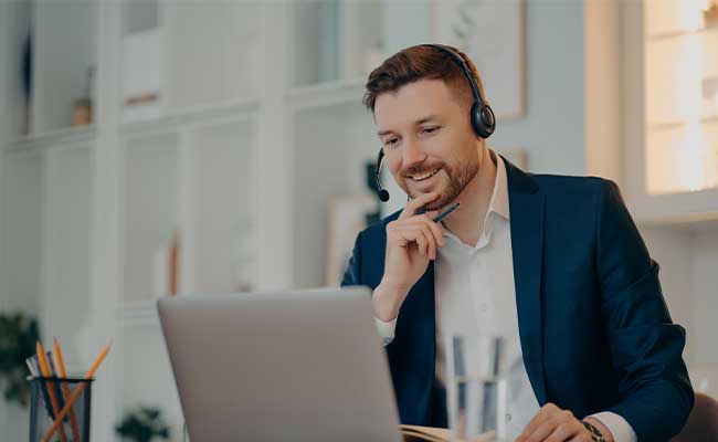 A smiling businessman wearing a headset sits at a desk during a video call on his laptop. He is dressed in a dark blue suit and holding a pen. The background shows a well-lit, modern office with shelves and decorative items.
