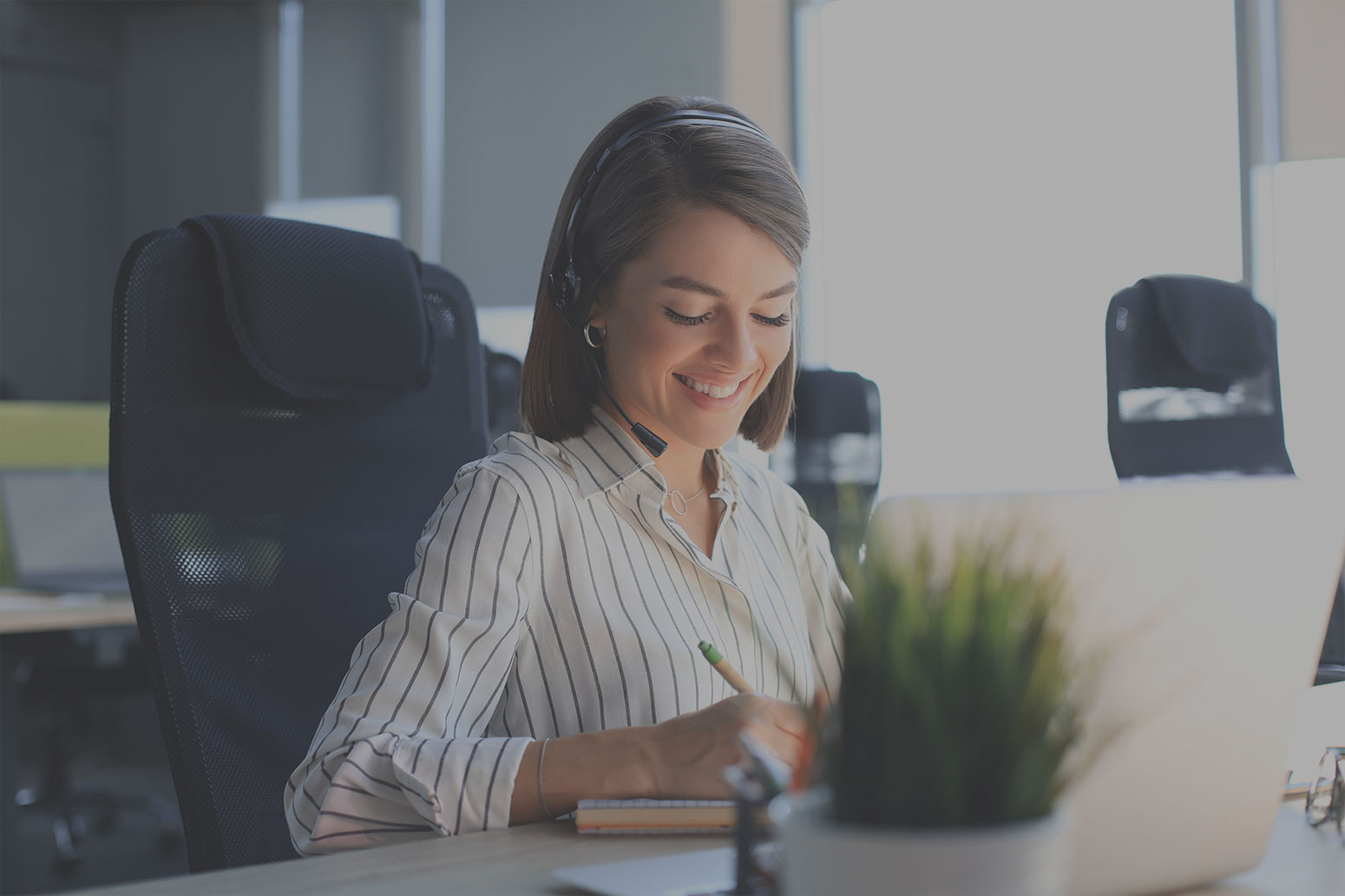 A smiling woman wearing a striped blouse and a headset is sitting at a desk in an office. She appears to be writing or taking notes while looking at something, possibly a laptop, in front of her. Office chairs and a plant are visible in the background.
