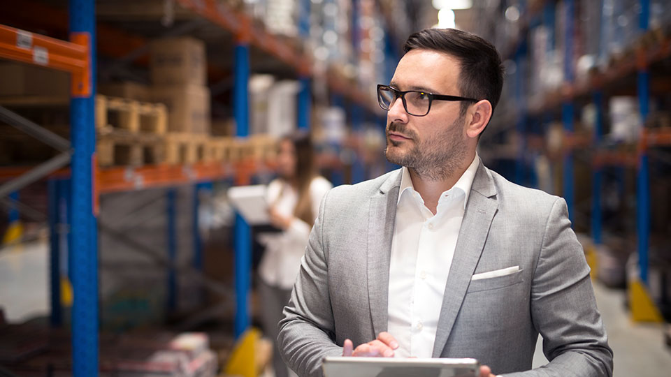 A man in a light grey suit with glasses is holding a tablet while inspecting inventory in a large warehouse. Shelves filled with boxes and another person working in the background suggest logistics or supply chain management.