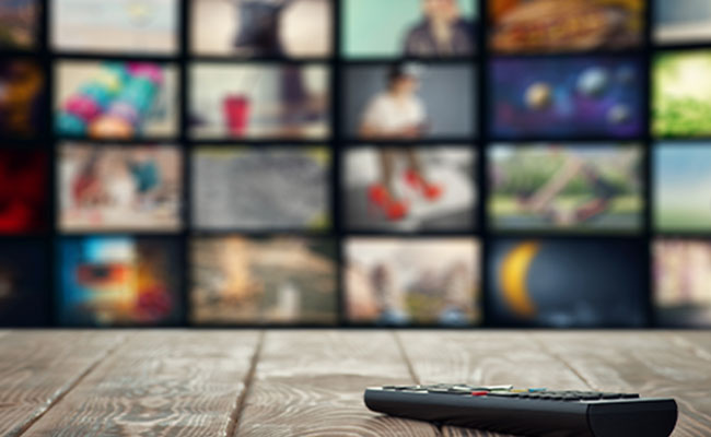 Remote control placed on a wooden table with a blurred background showing multiple TV screens.