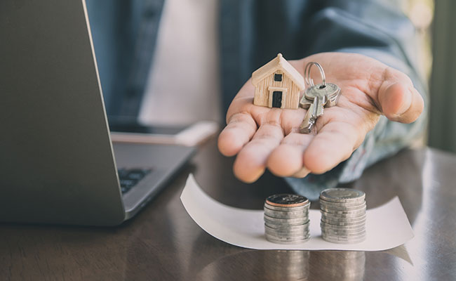 Person holding a small wooden house model and keys in one hand, with coins stacked on paper and a laptop on the table.