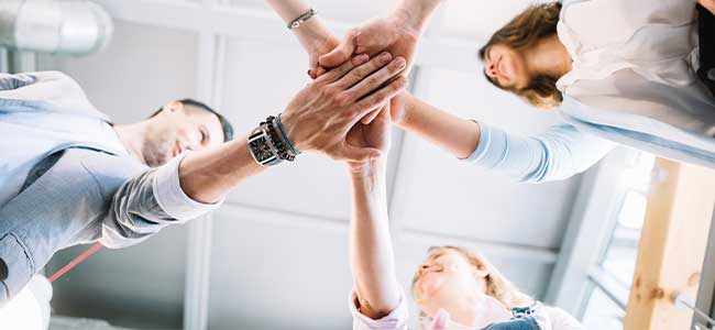 Four team members stacking their hands together in a gesture of unity and teamwork, viewed from below.