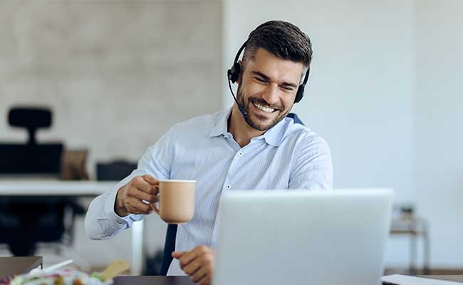 Smiling man with a headset holding a coffee mug while having a video call on a laptop in a modern office.