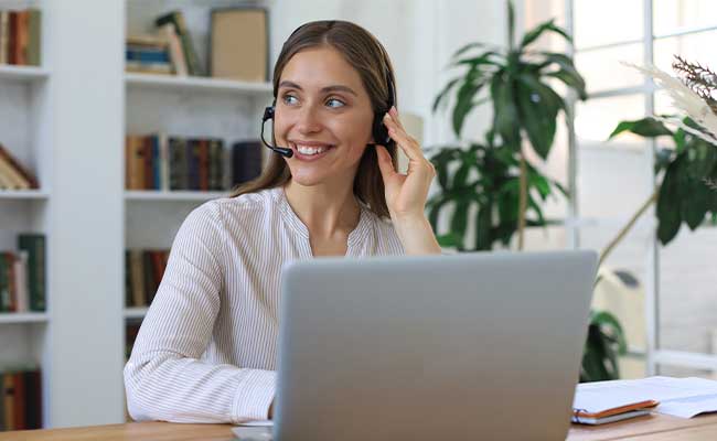 Smiling woman wearing a headset, sitting at a desk with a laptop, in a bright room with bookshelves and plants in the background.