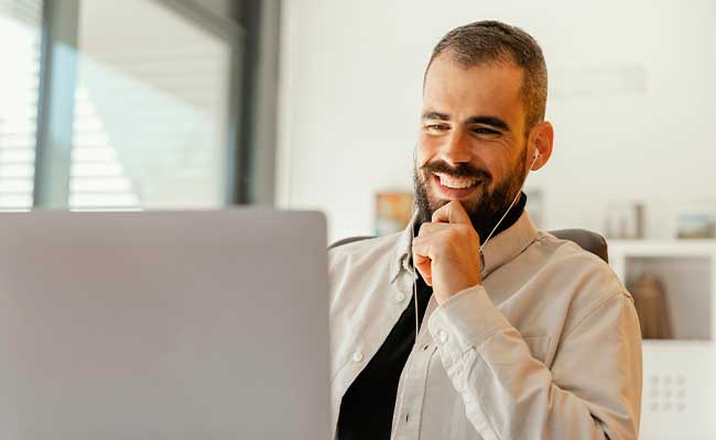 A smiling man with earbuds sitting at a desk and looking at a laptop screen during a video call or online meeting.