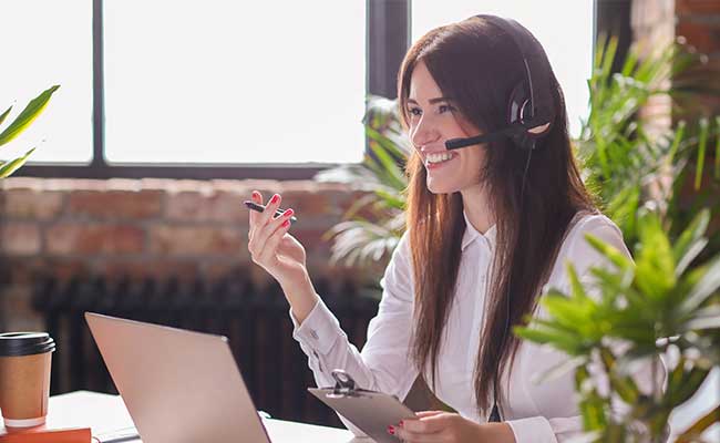 Smiling woman wearing a headset having an online meeting at her desk, holding a clipboard and pen.