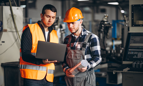 Two industrial workers discussing machine operations using a laptop in a production environment.