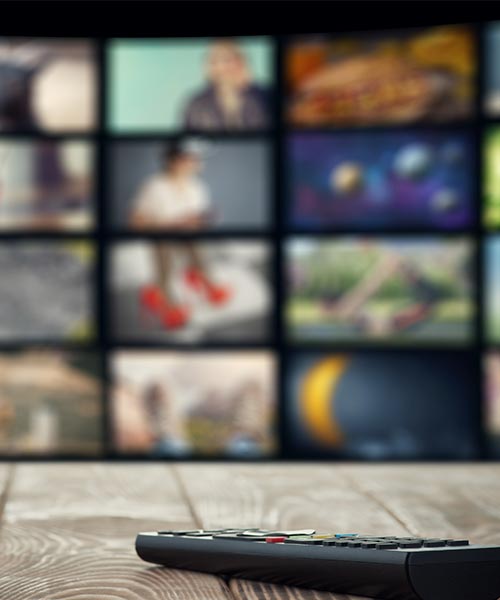 A remote control placed on a wooden table with a blurred background showing multiple TV screens.