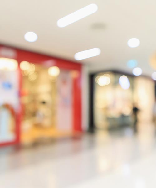 Blurred image of a modern shopping mall with storefronts and illuminated ceiling lights.