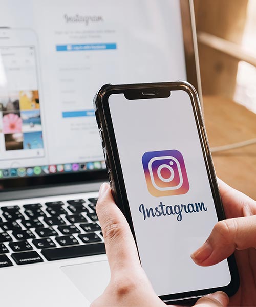 A person holding a smartphone showing the Instagram app login screen, with a laptop in the background.