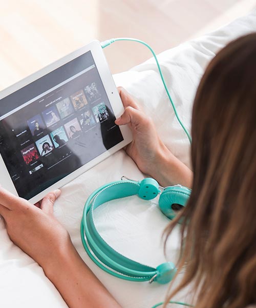 A woman using a tablet to browse music streaming content, with teal headphones next to her.